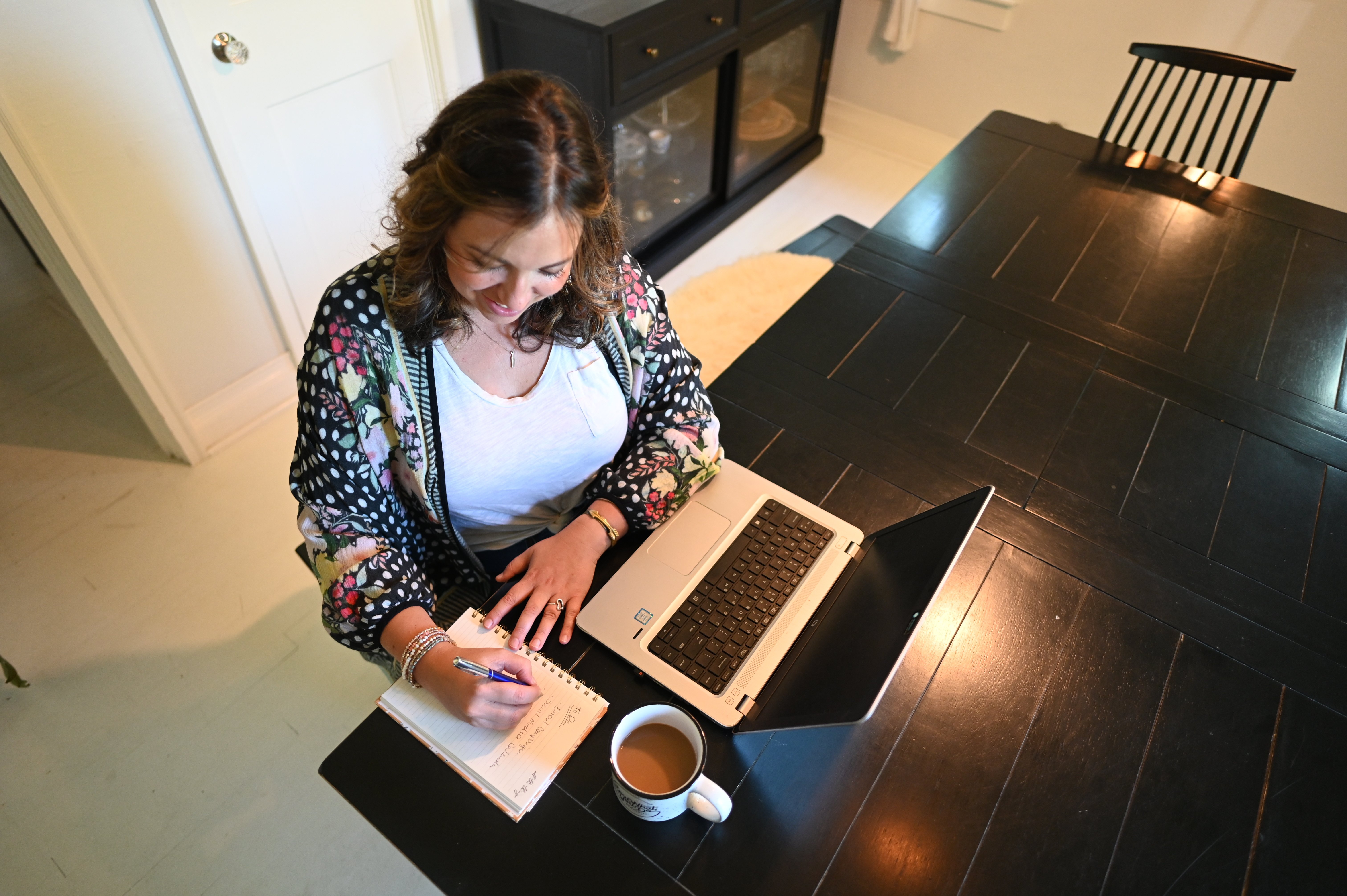 Woman sitting at a table, with a computer and coffee, writing in a journal